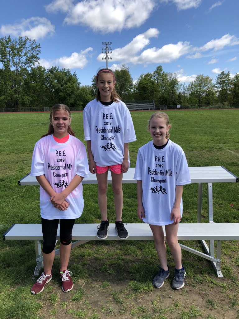 Three fifth-grade girls stand wearing their white presidential mile t shirts. the first place winner is in center, standing highers than the others.