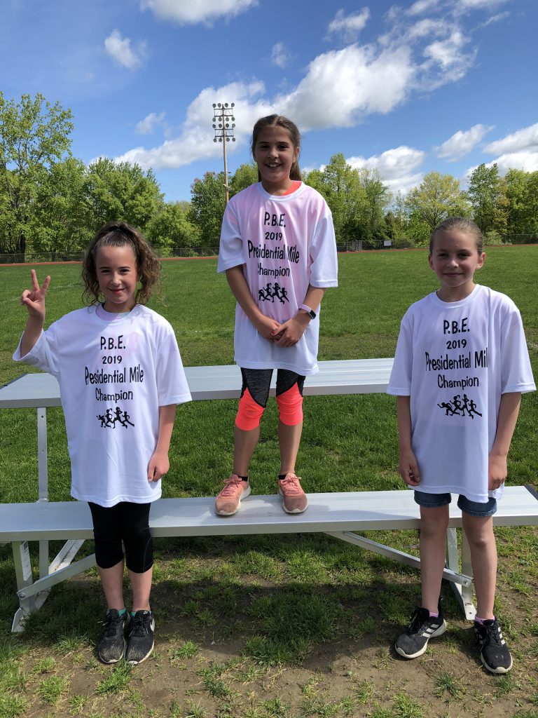 Three fourth-grade girls wearing white presidential mile t shirts. Winner in center stands higher. The girl on the left is holding up her index figure to show they are number 1.