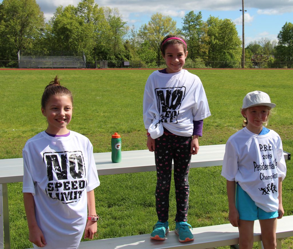 Three third-grade girls dressed in white t-shirts noting the presidential mile. First place winner in center standing higher than the others 