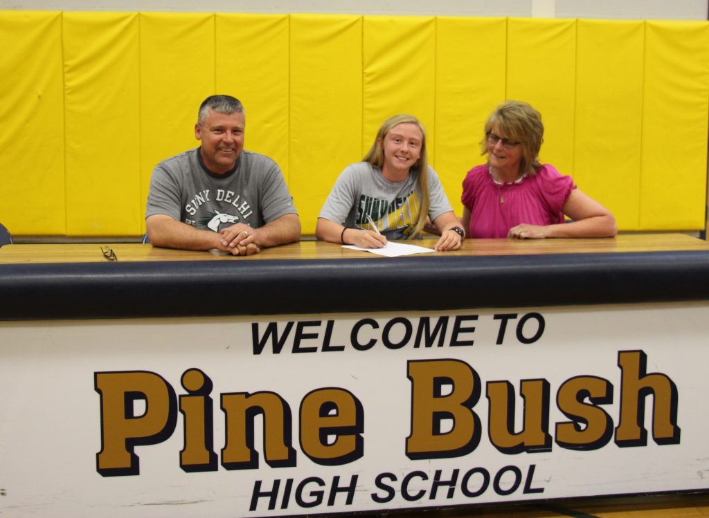 A young woman with blonde hair wearing a gray shirt sits at a large table with a sign "Welcome to Pine Bush High School" on front. Her parents are on either side of her as she signs a letter.