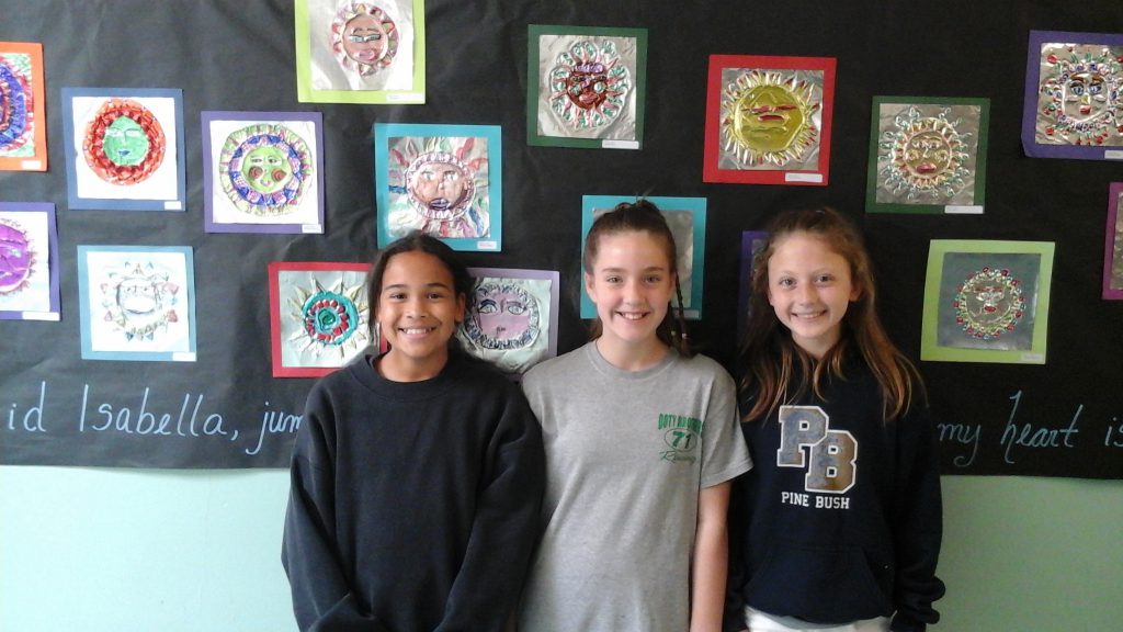 Three older elementary school girls stand in front of a wall of art.