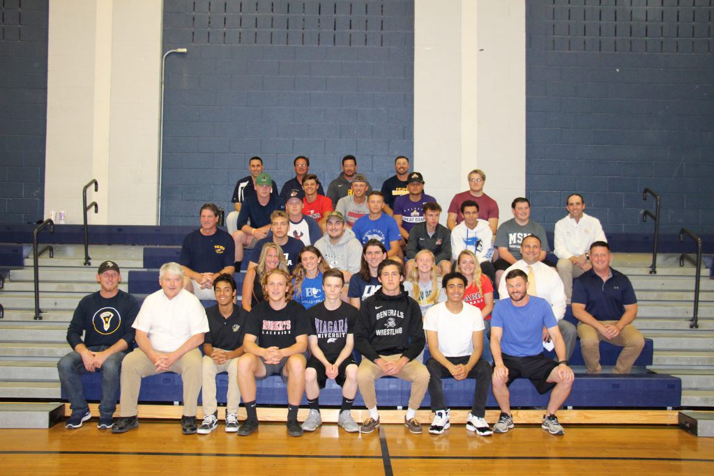 Twenty-one high school senior athletes sit on bleachers with their coaches