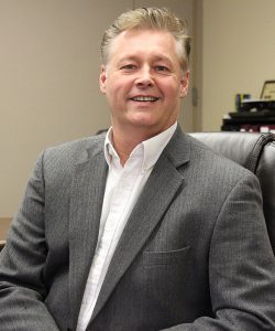 A man with blonde hair sitting in an office chair wearing a white button down shirt and gray suit jacket.