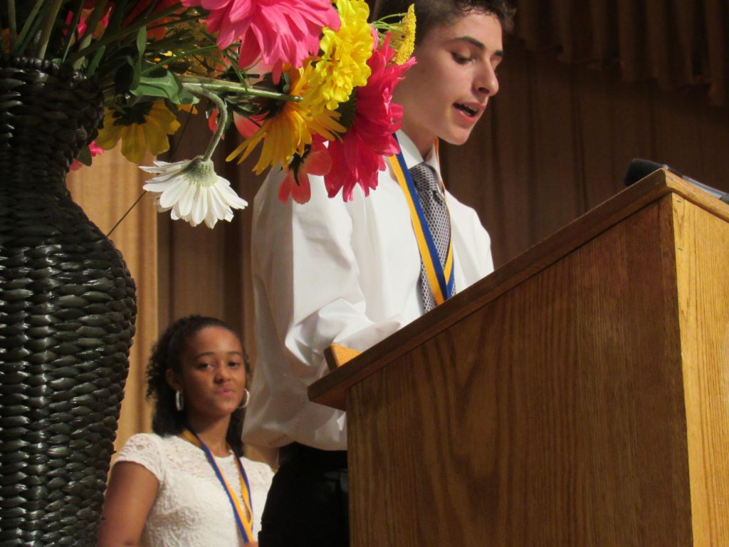 A young man speaking at a podium in a white shirt and tie wit a young woman standing behind in a white dress. Flowers are off to the side.