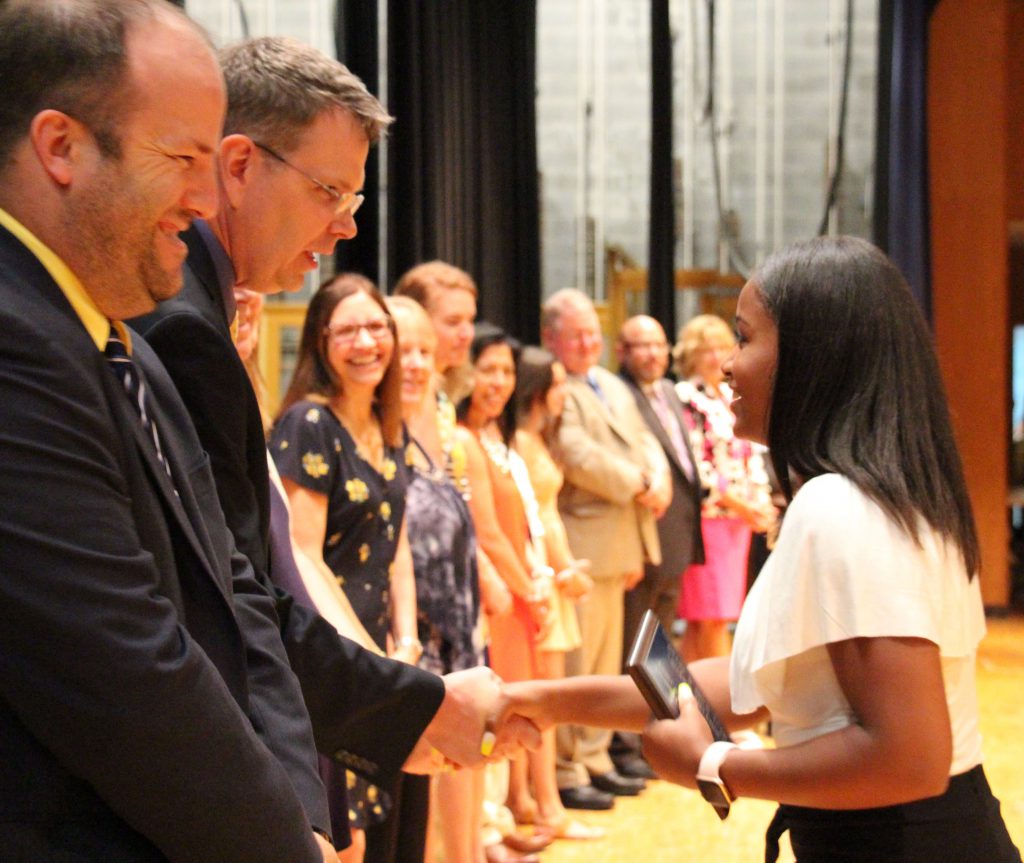 A student dressed in a white blouse and black pants shakes hands with her teachers, who are in a line