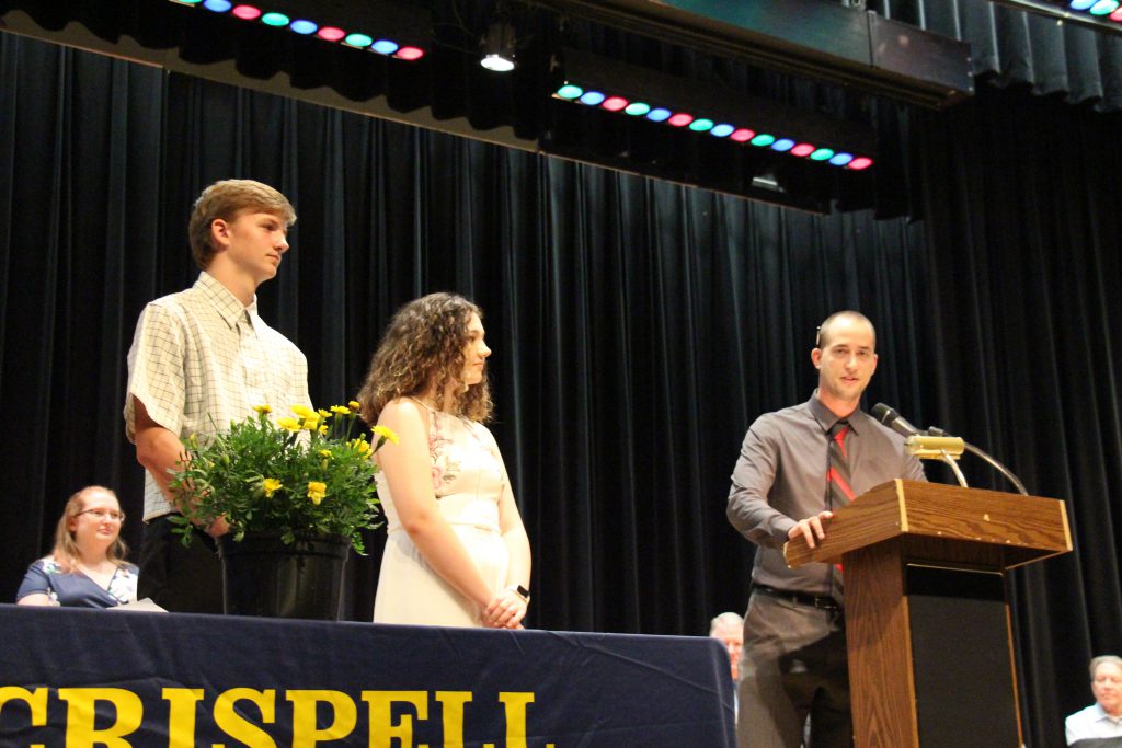 A young man and young woman stand and listen while a man speaks at the podium before presenting them with awards