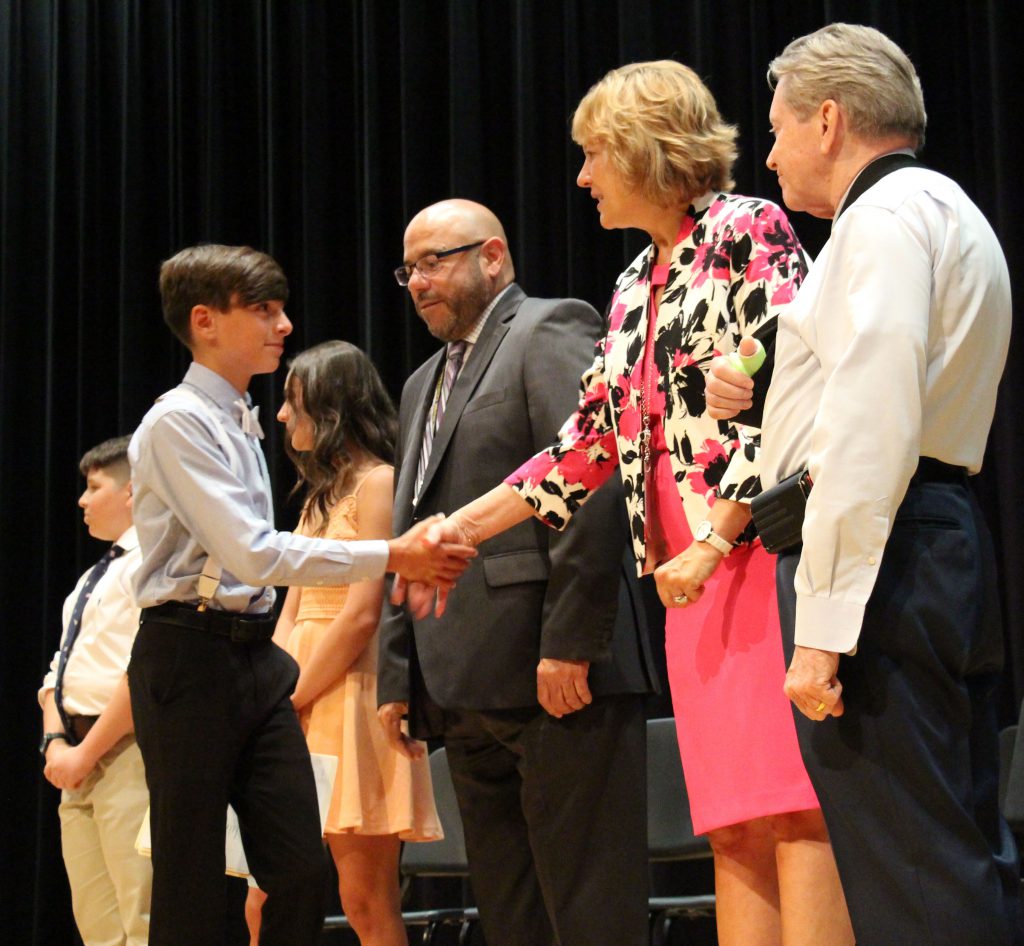 A young man in a shirt and tie shakes hands with a woman in a pink dress as other administrators look on