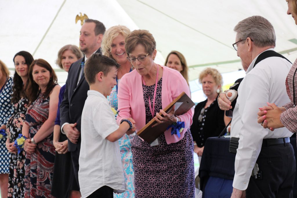 A boy in a white shirt shakes hands with a woman in a dress and pink sweater as he receives a plaque.