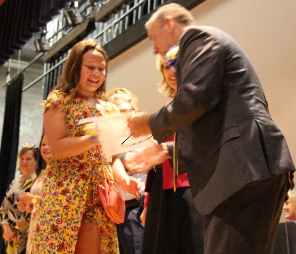 A fifth-grade girl in a yellow dress smiles as she accepts an award from a her principal, a man in a gray suit