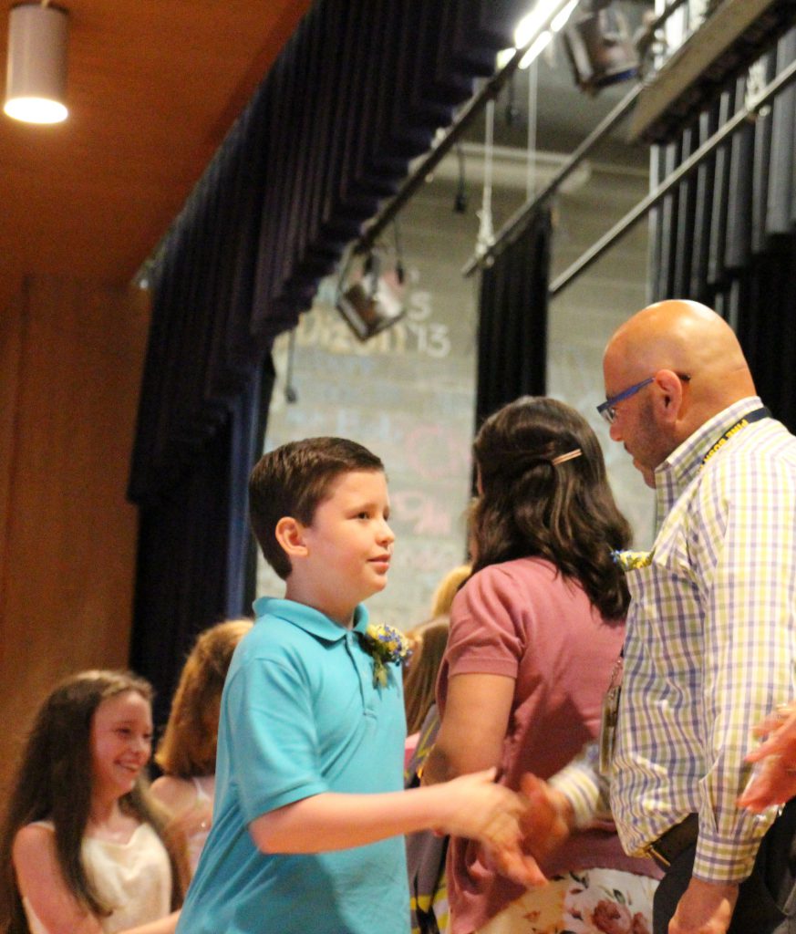 A young man in an aqua colored polo shirt shakes hands with a man in a plaid button down shirt and glasses.