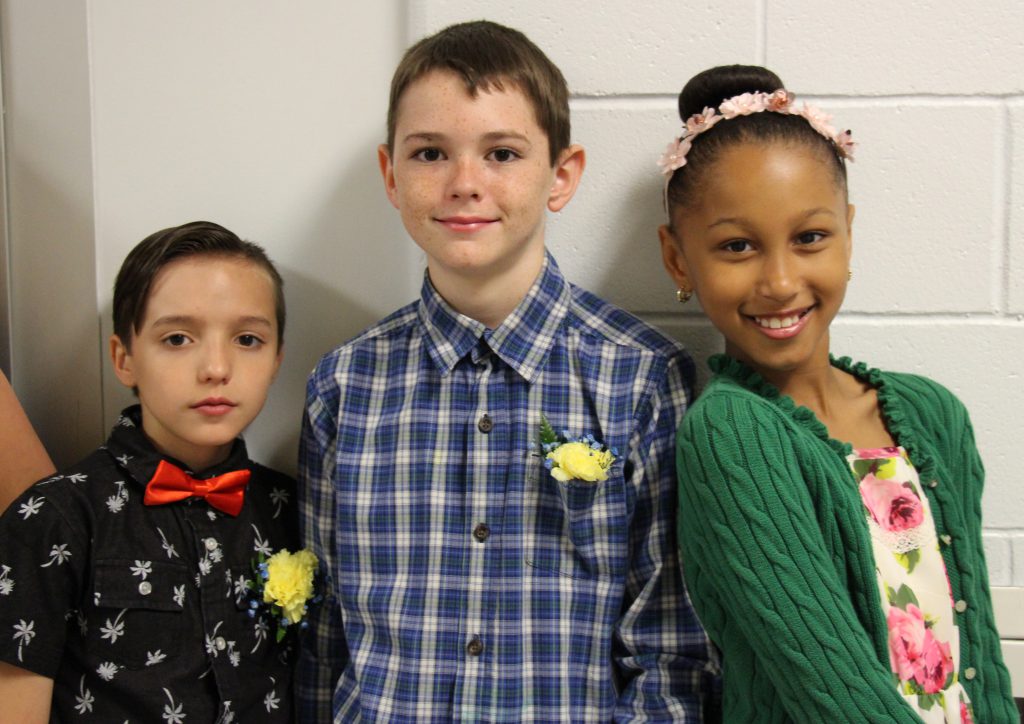 Three fifth-grade students stand against a wall, On left is a boy in a black shirt and red bow tie, boy in center is wearing a plaid shirt, girl on right has her hair in a bun and is wearing a green sweater. They're smiling and wearing flowers pinned to their shirts.