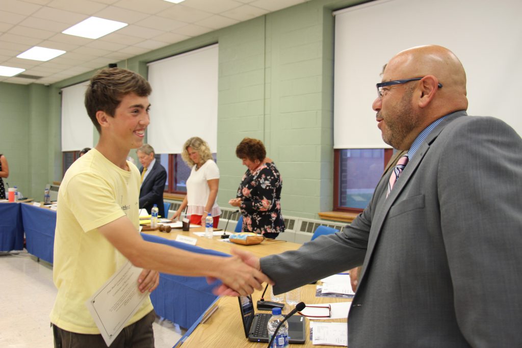 A young man in a yellow shirt shakes hands with a man wearing glasses and a gray suit