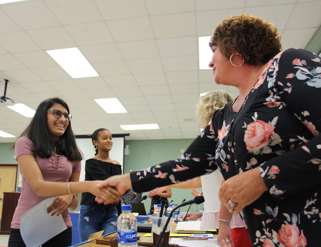 A young woman with long dark hair and glasses smiles as she shakes hands with a woman with short brown hair wearing a flowered dress