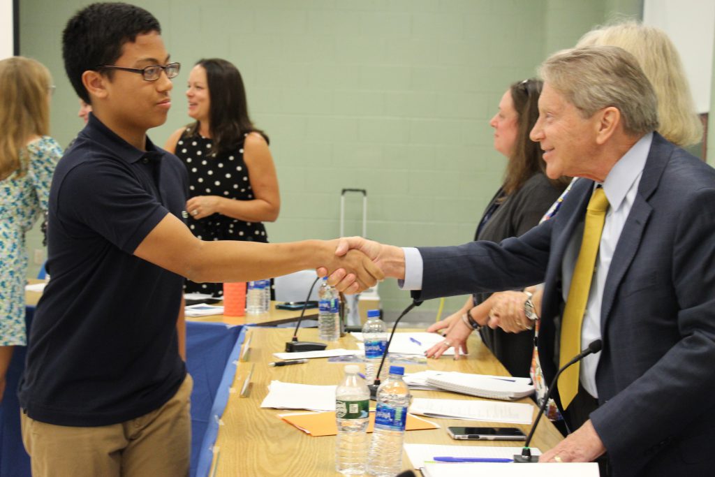 A young man wearing a black shirt and glasses shakes hands with a man wearing a blue suit and gold tie