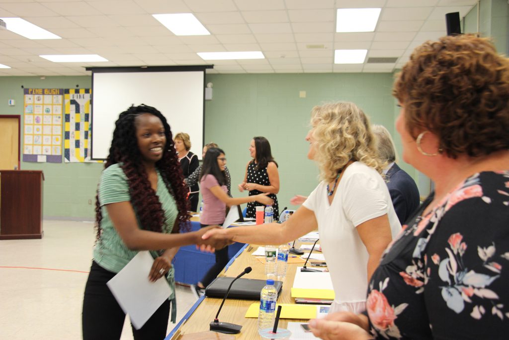 A young woman with long wavy dark hair smiles and shakes hands with a woman with blonde hair wearing a white shirt