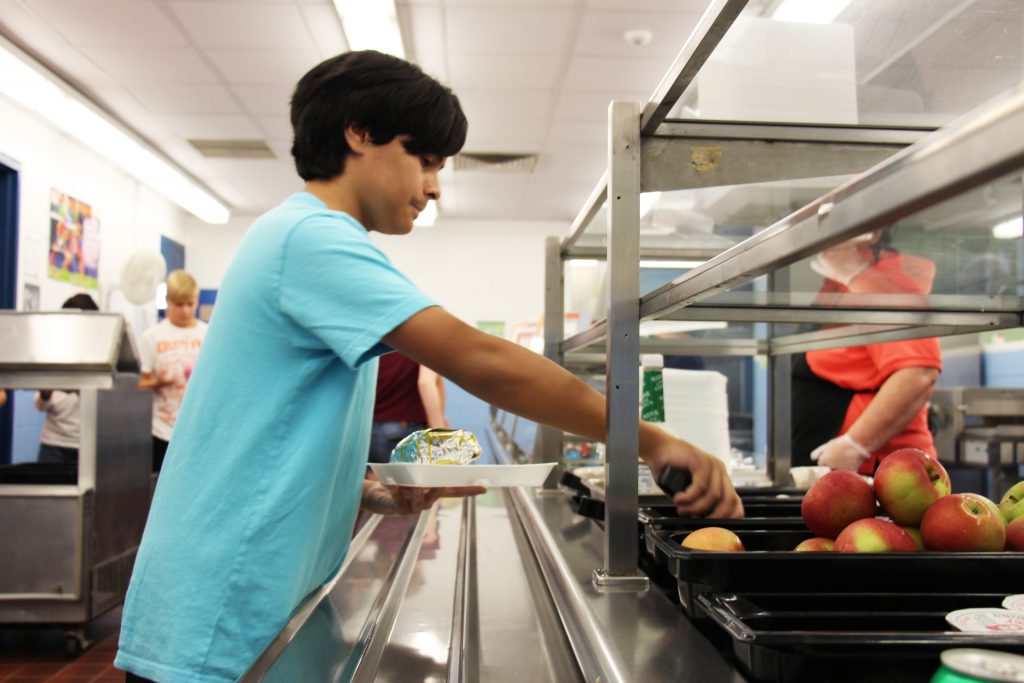 A middle school boy with dark hair wearing a bright blue t-shirt. He is on the cafeteria line reaching in with tongs. Apples are piled in a bin.