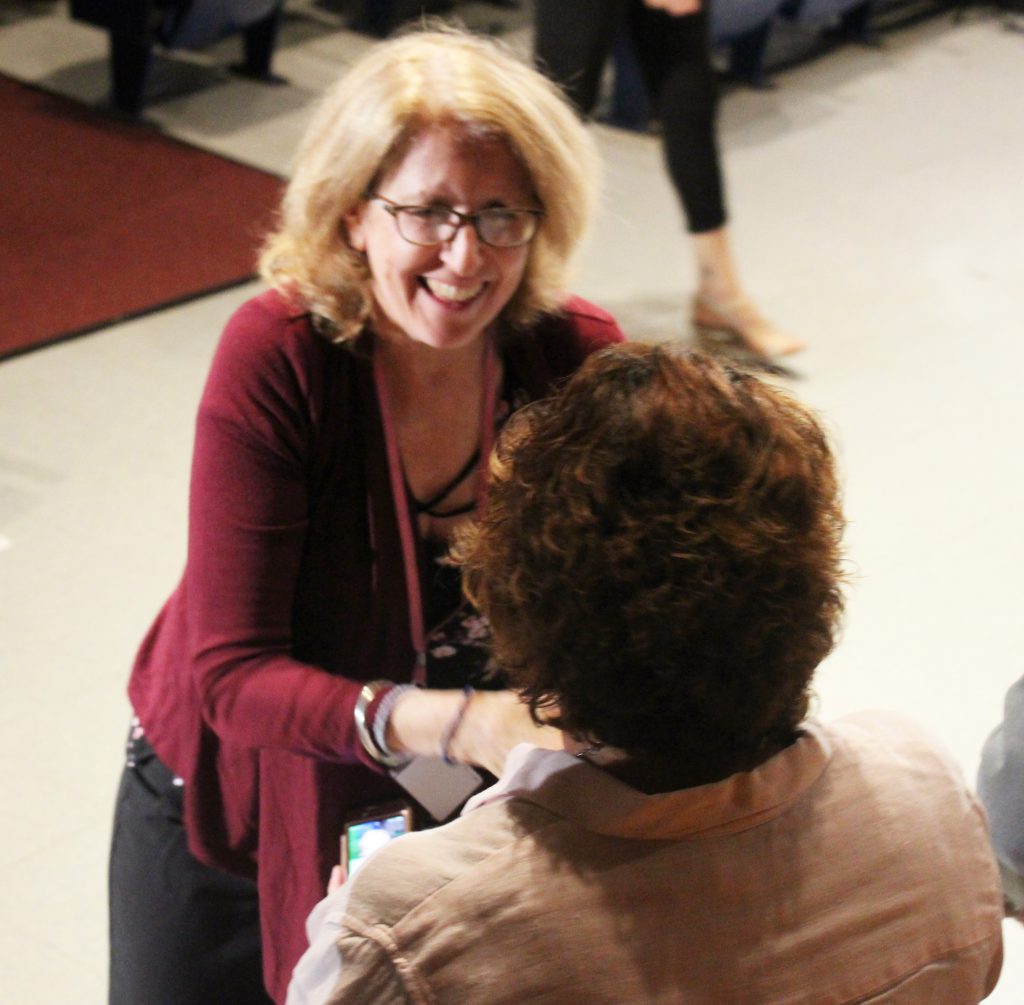 A woman with blonde hair and glasses wearing a red blouse smiles and shakes hands with another woman.