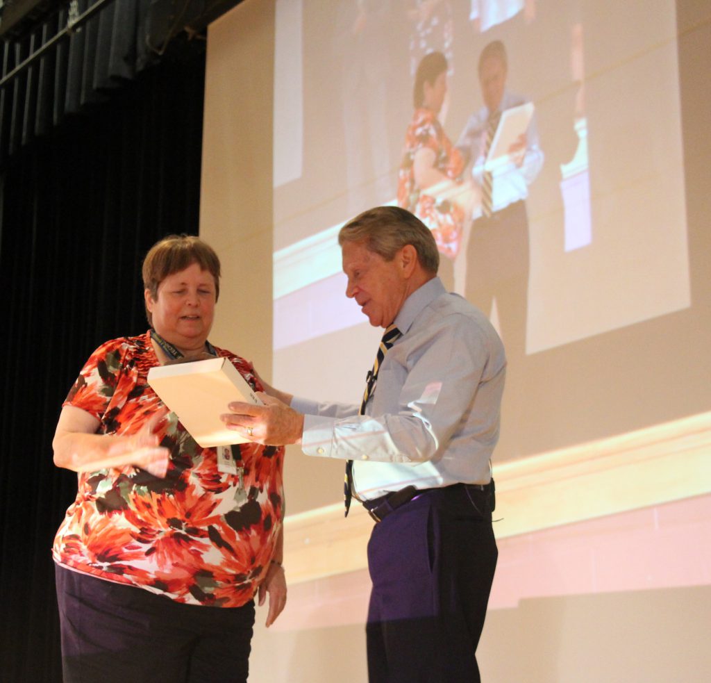 A woman with short hair accepts an award from a man wearing a blue dress shirt and tie. There is a large screen in the background showing the two.
