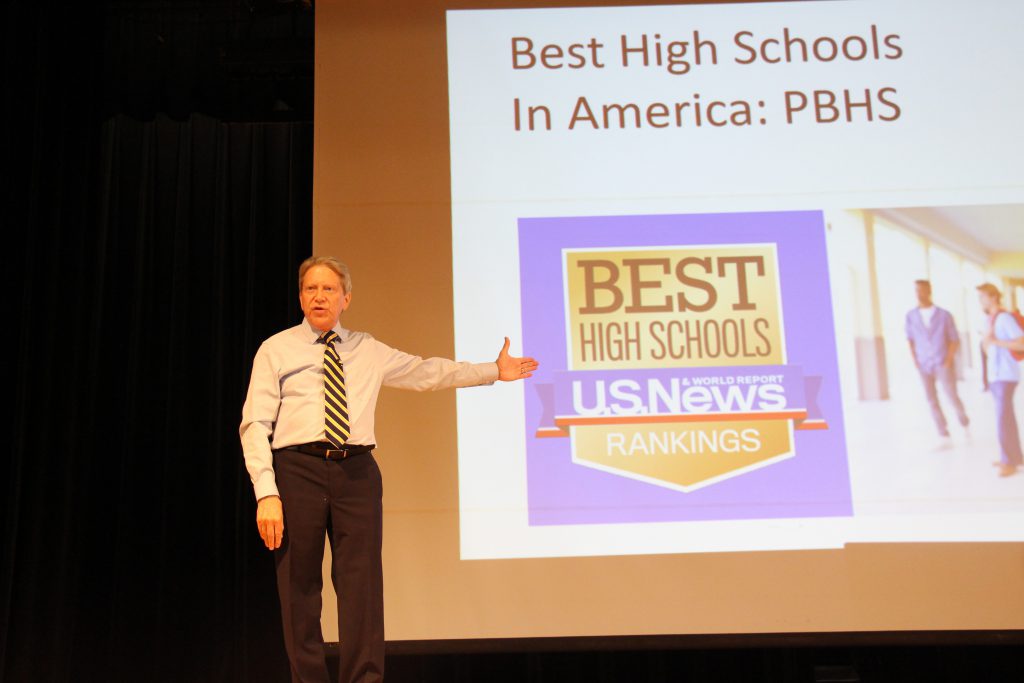 A man wearing a shirt and tie standing in front of a large projection screen holds his hand up pointing to the screen showing Pine Bush High School was named a best high school by U.S.. News and World Report