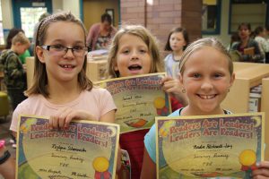 Three elementary students smile as they hold up their reading certificates that say Readers Are Leaders! Leaders are Readers!