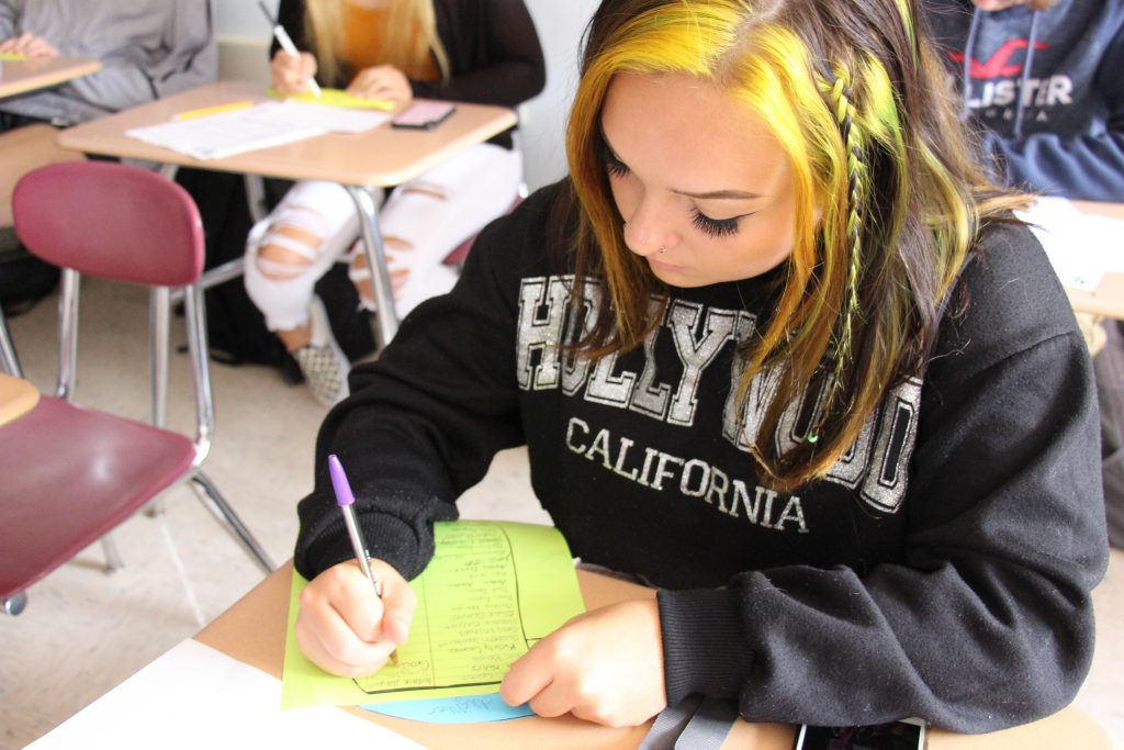 A high school senior girl wearing a Hollywood California sweatshirt sits at a desk and writes on a piece of paper