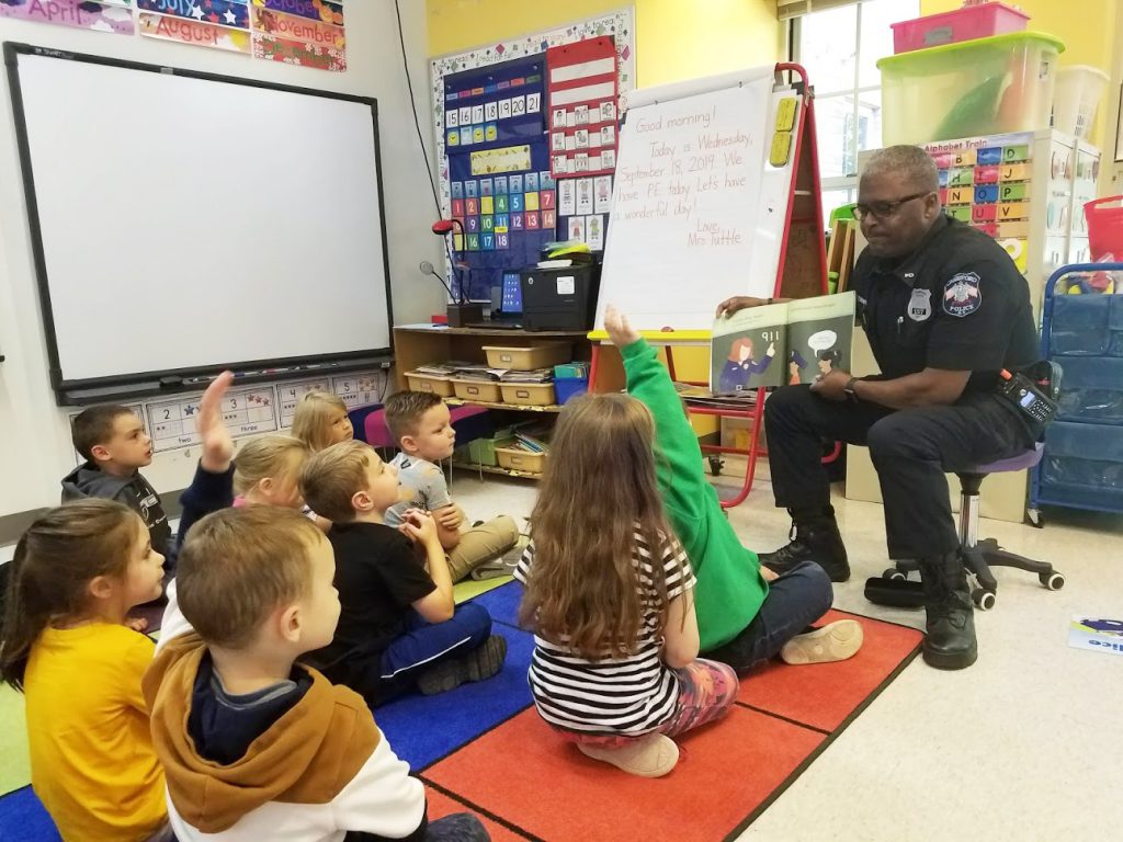 A police officer is sitting in front of the class with a book opened toward the students who are facing him, sitting on a rug. Some students have their hands up