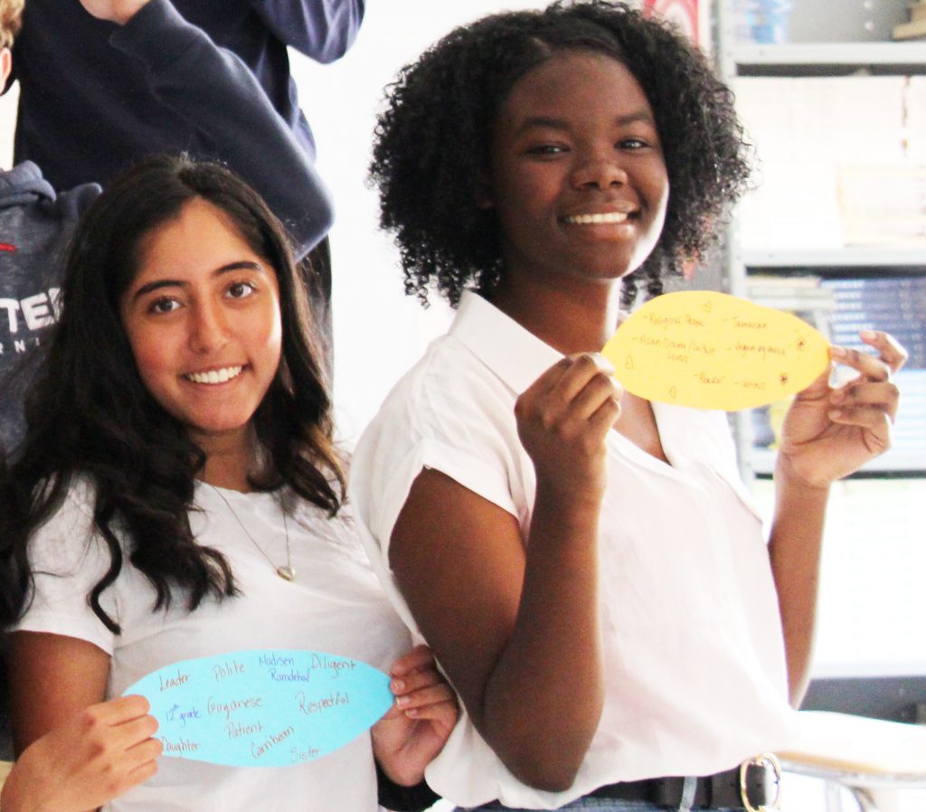 Two young ladies, both wearing white shirts, hold their leafs up and smile