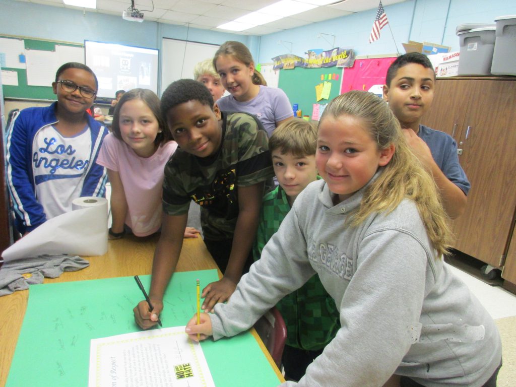 A group of seven middle school students stand around a table and sign a poster that has the resolution of respect