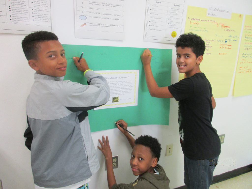 Three young men sign a poster that hangs on a wall.