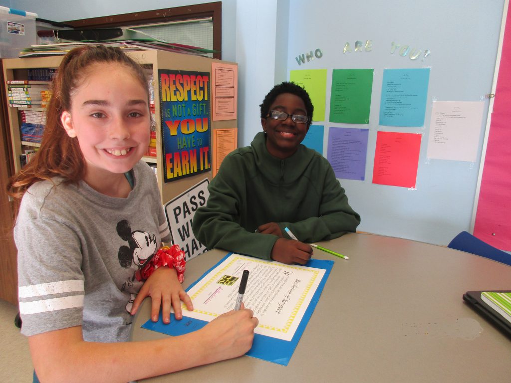 Two students sign poster that sits on a desk