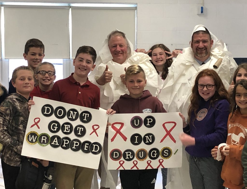 Seven middle school students gather around their principal and assistant principal who are wrapped up in toilet paper. Two students hold signs that say Don't Get Wrapped Up In Drugs"