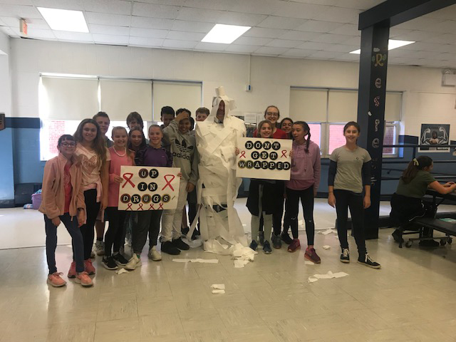Twelve middle school students stand with their principal who is wrapped from head to toe in toilet paper. Two students are holdings signs that say "Don't Get Wrapped Up In Drugs"
