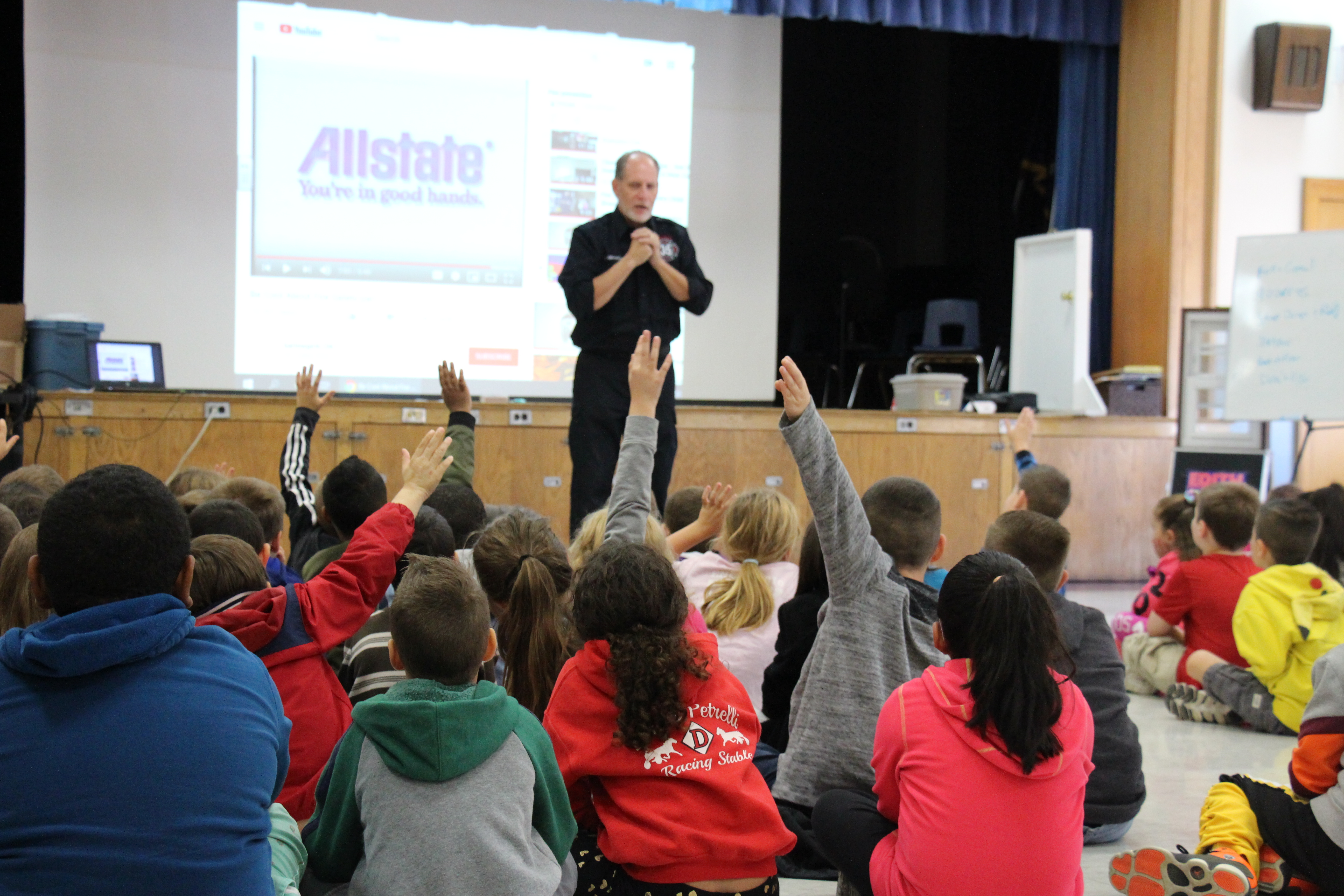 A firefighter stands in front of a large group of students, some of whom have their hands up to answer questions.