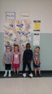 Four Pre-K students stand in front of the paper tree they created using their handprints with their promises on them.