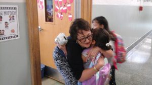 A female teacher wearing glasses bends down and gives a hug to a young elementary school girl