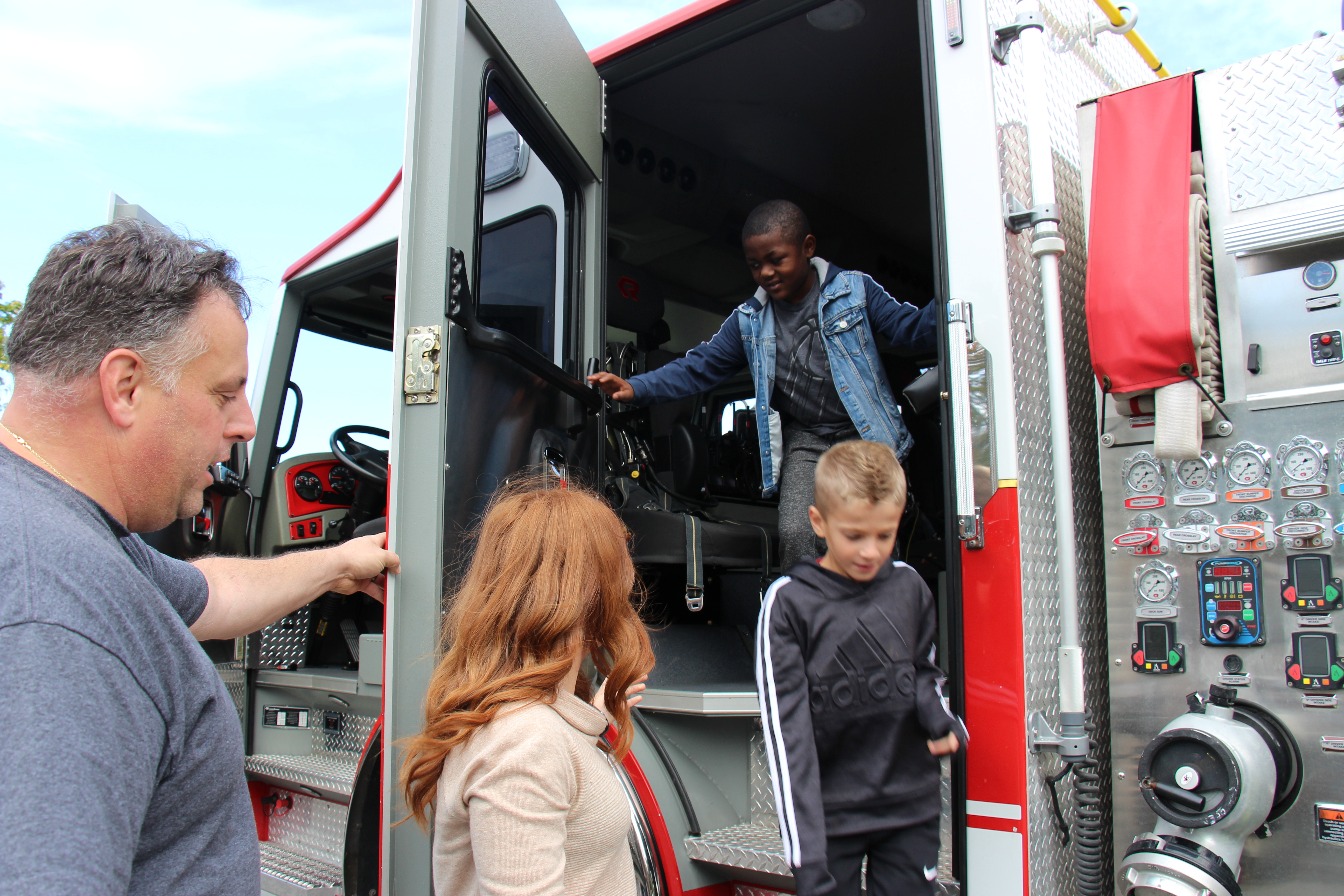 Two elementary school students come down the steps of a fire engine as a man holds the door open and a woman helps them.