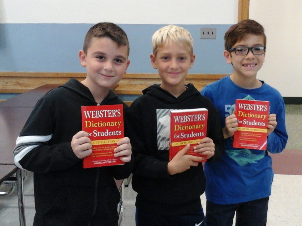 Three third-grade boys smile as they hold up their red dictionaries