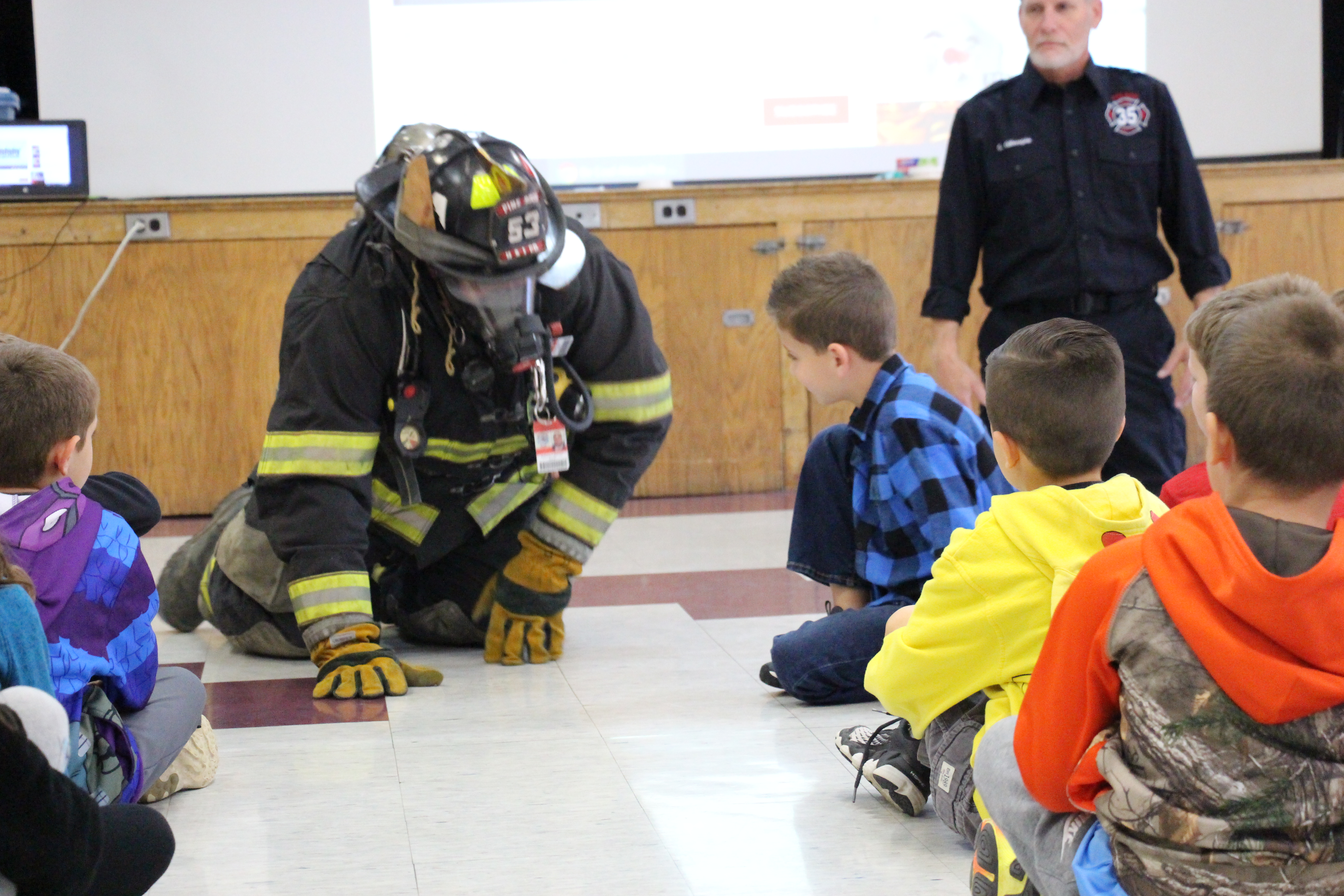 A man in full firefighter gear is on his hands and knees about to crawl through a group of elementary school students.