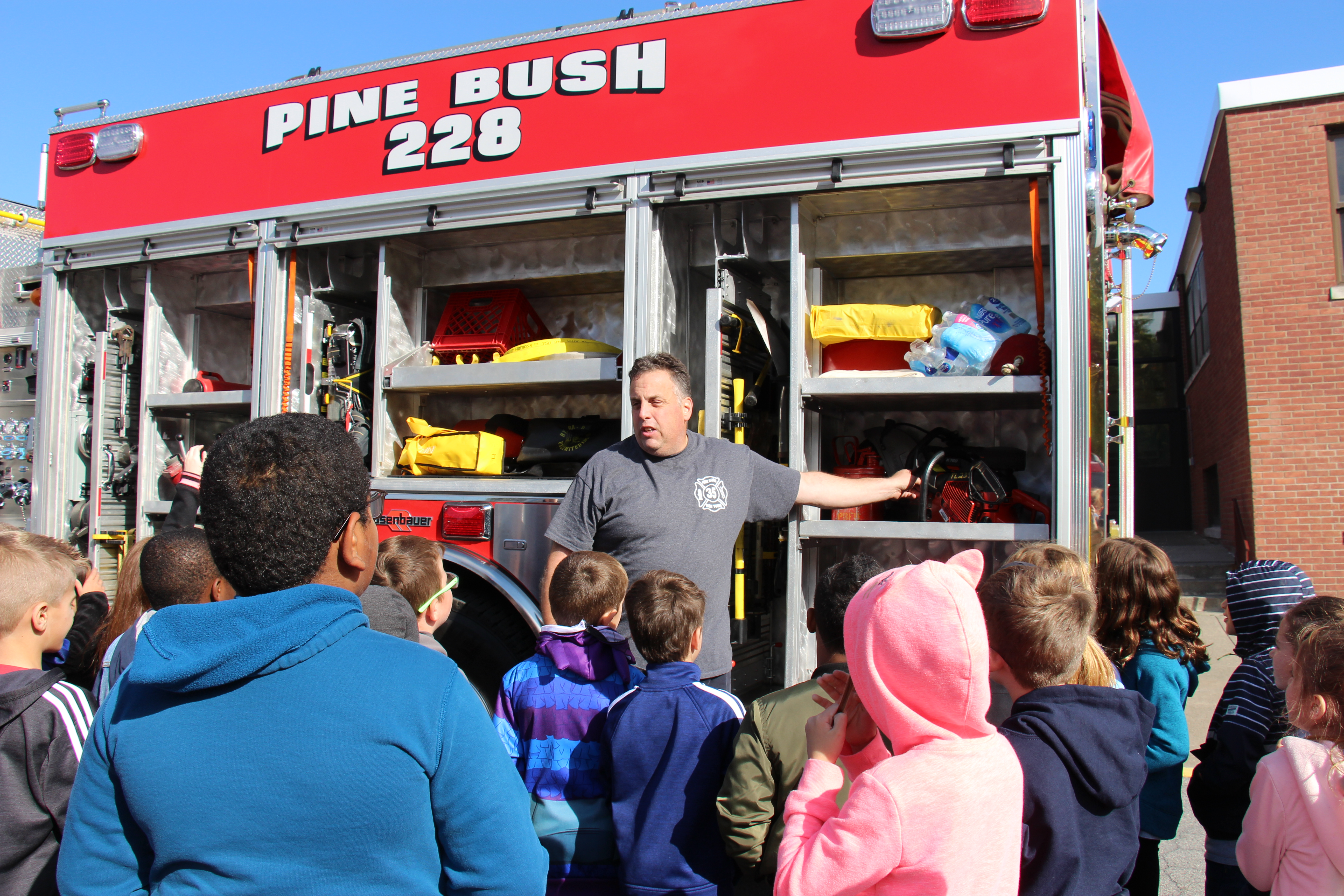 A large group of elementary students listen as a man stands in front of a fire engine pointing out all of its equipment during fire safety week.