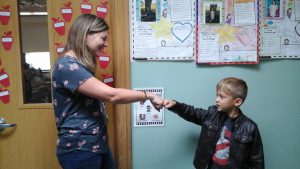 A female teacher standing in a doorway fist bumps a young male student