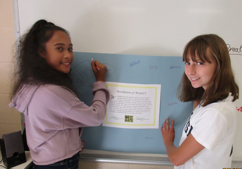Two young women smile as they sign a poster on a wall