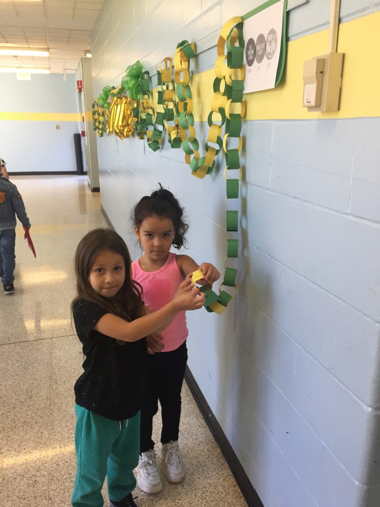 Two elementary school age girls hold a chain made of paper links, most of which is on the wall