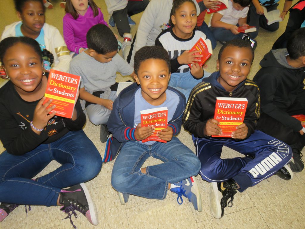 Three students sitting on the floor smile while holding up their new red dictionaries.