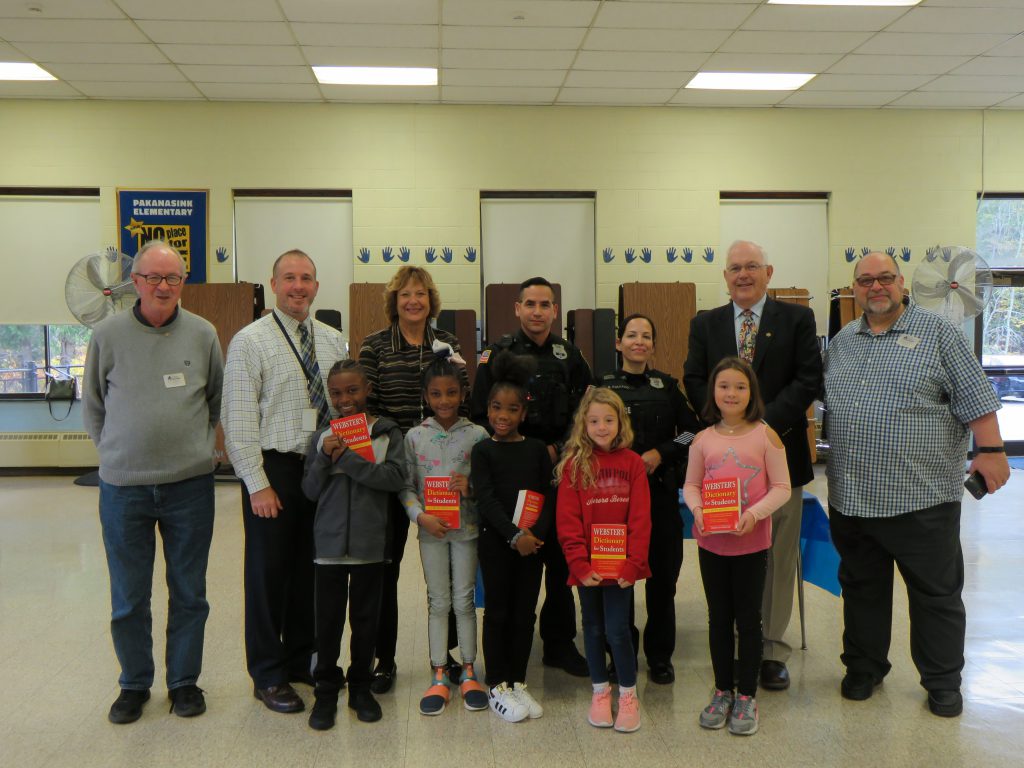 Seven adults stand behind five third-grade students who are all holding their new red dictionaries.