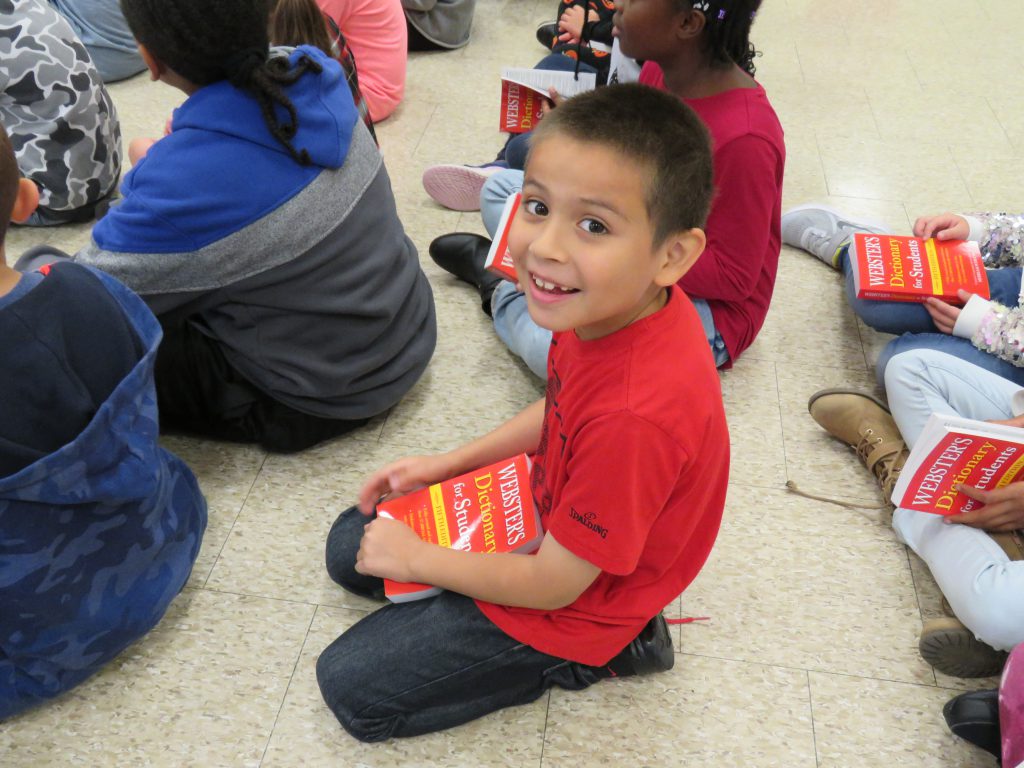 A third-grade boy with short dark hair wearing a red t shirt smiles as he holds his new red dictionary.