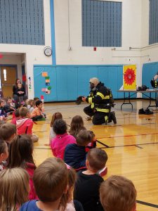 A firefighter puts on his gear in front of a large group of kindergarten and pre-k students