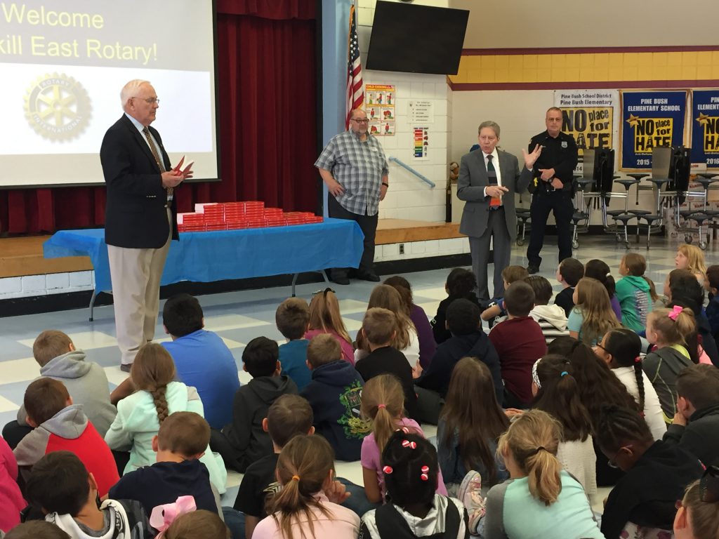 A large group of third-grade students sit on the floor and listen to a man in a suit talk to them. On the table behind them is a large stack of red dictionaries. Other adults look on.