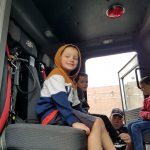 A young boy wearing a hooded sweatshirt sits in a fire truck and smiles. Other boys are also in their seats smiling.