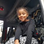 A little girl dressed in a black shirt and black and white pants smiles from inside the fire truck.