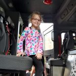 A girl wearing a multi-colored shirt and glasses smiles from inside the fire truck.